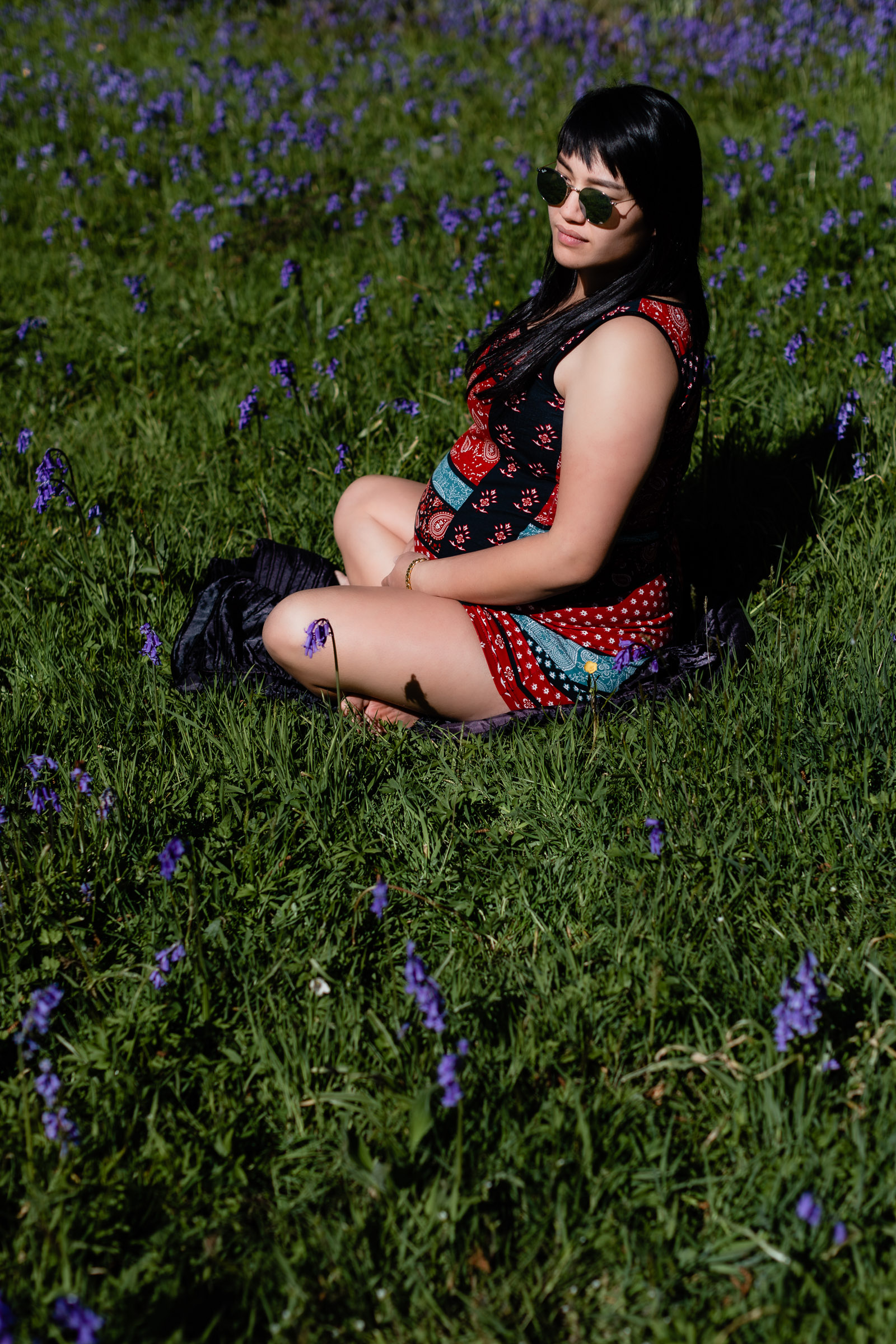 A young woman in late pregnancy sitting cross legged on a grassy area with bluebells on a maternity shoot in West Sussex