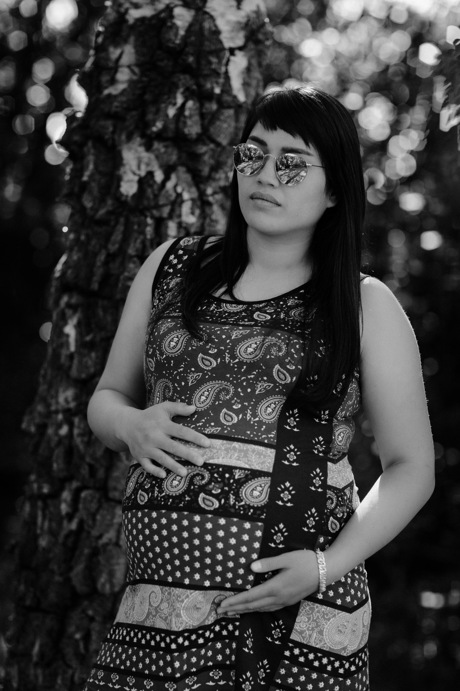 A black and white image of a pregnant young woman wearing sunglasses and standing under a tree on a maternity shoot in Horsham West Sussex