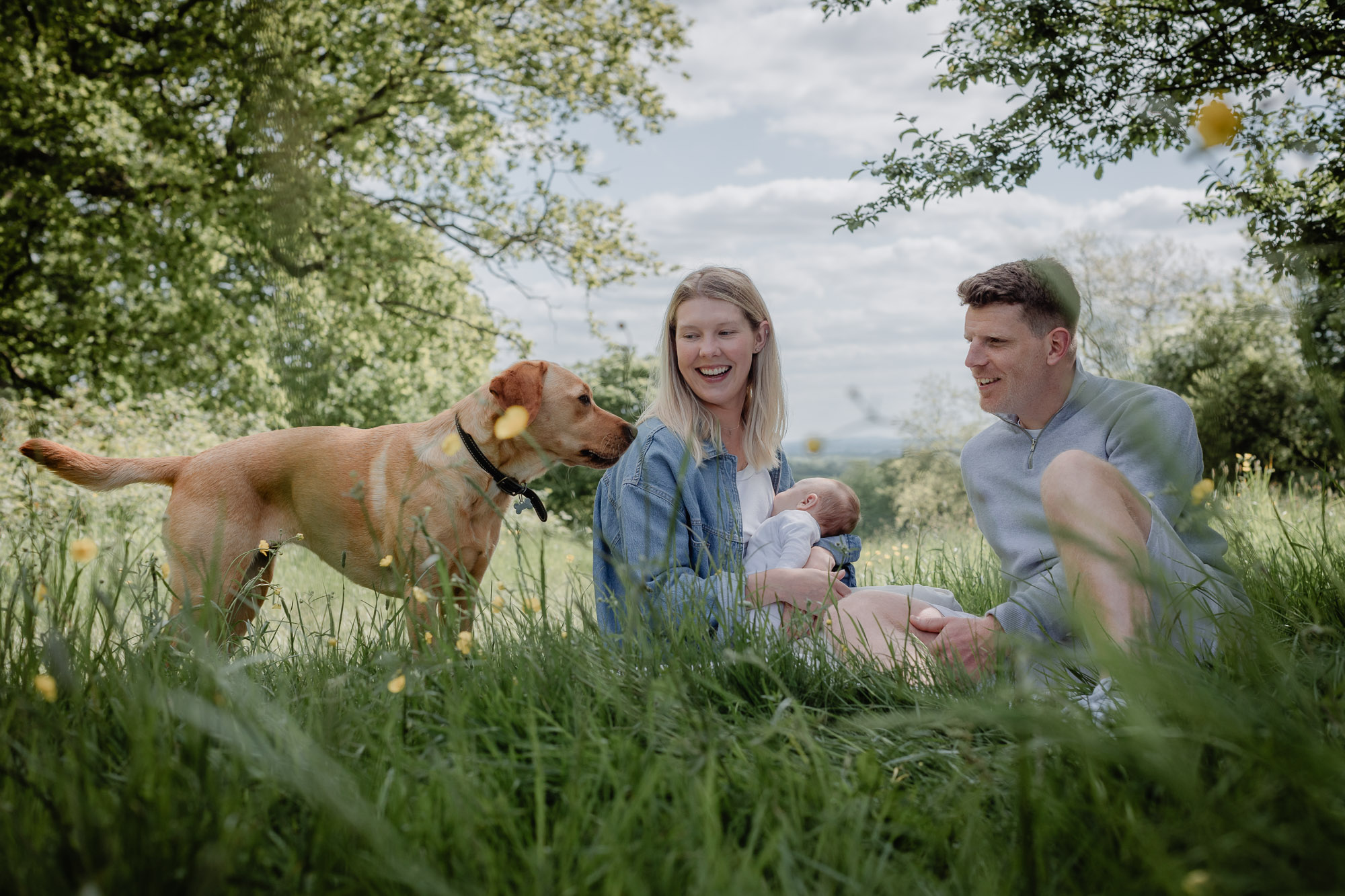 A young couple sitting on the grass with the mother nursing a newborn baby and both smiling at their Labrador, who stands besides them, on a newborn shoot in Horsham, West Sussex.