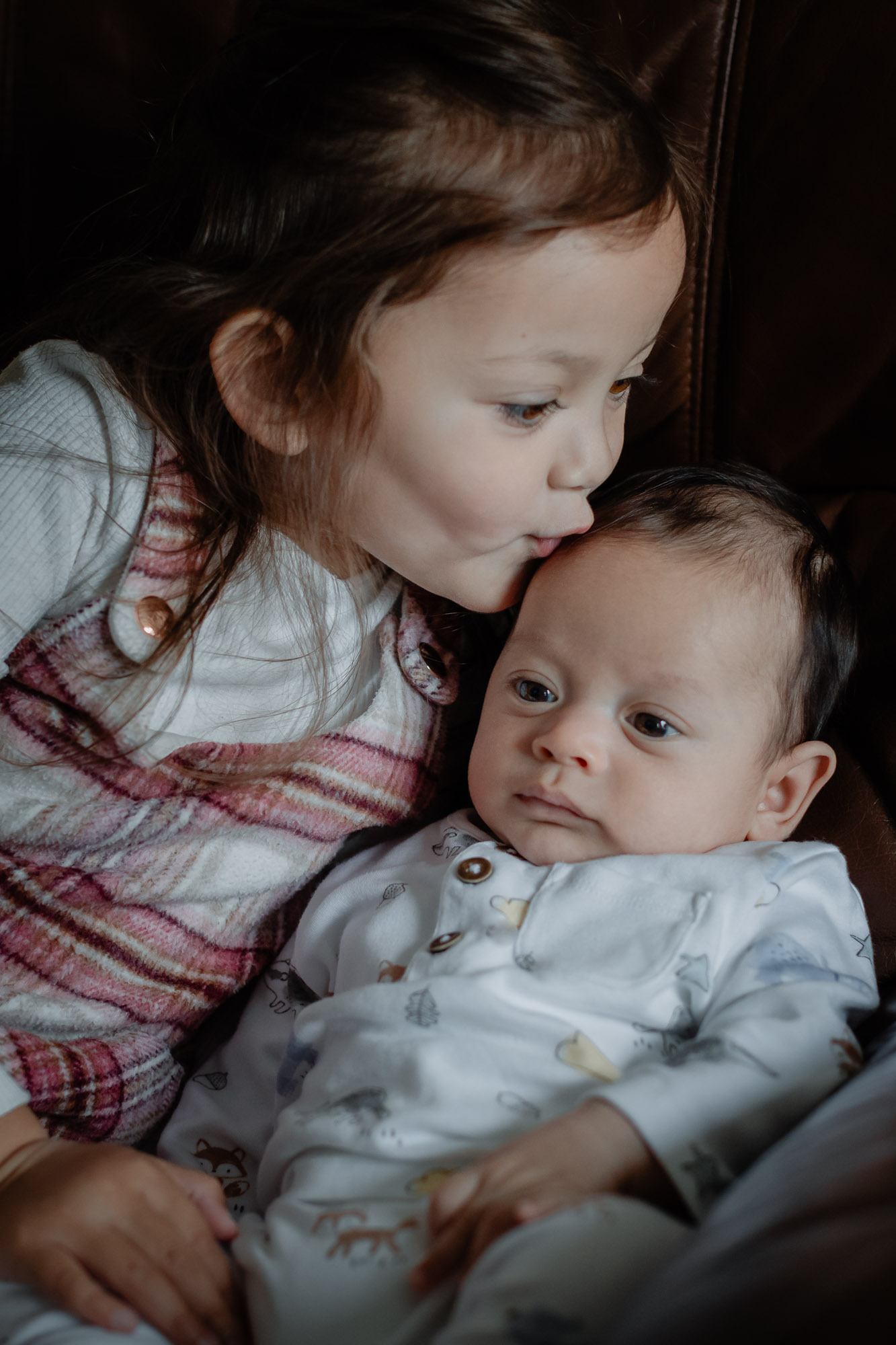 A little girl cuddling up to her baby brother and leaning over to kiss his head on a baby and family photoshoot in Horsham, West Sussex.