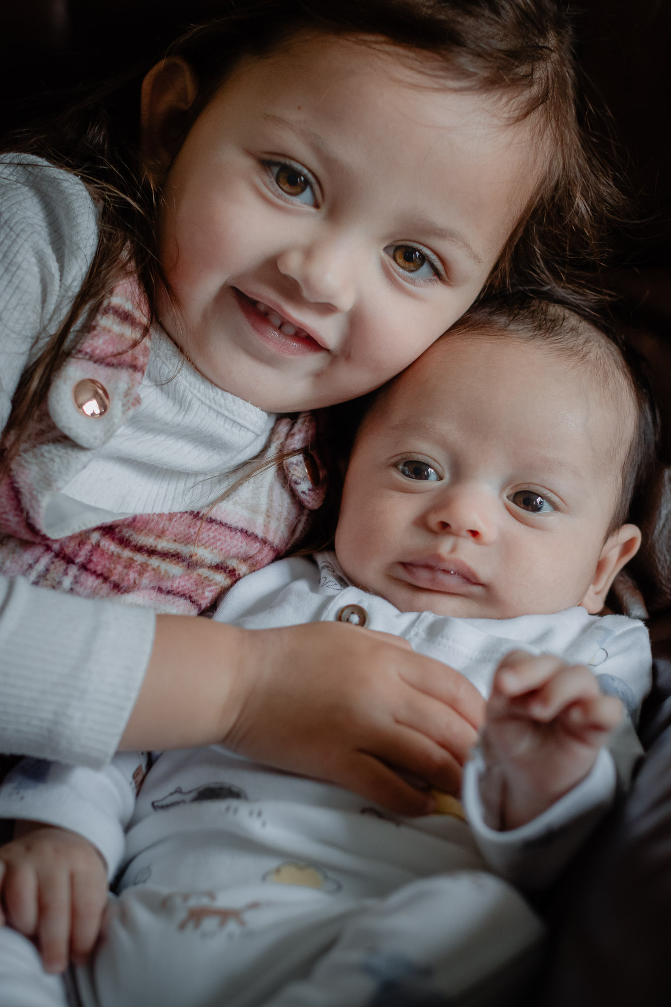 A little girl cuddled on a sofa with her baby brother, looking towards the camera and smiling on a baby photoshoot in Horsham, West Sussex.