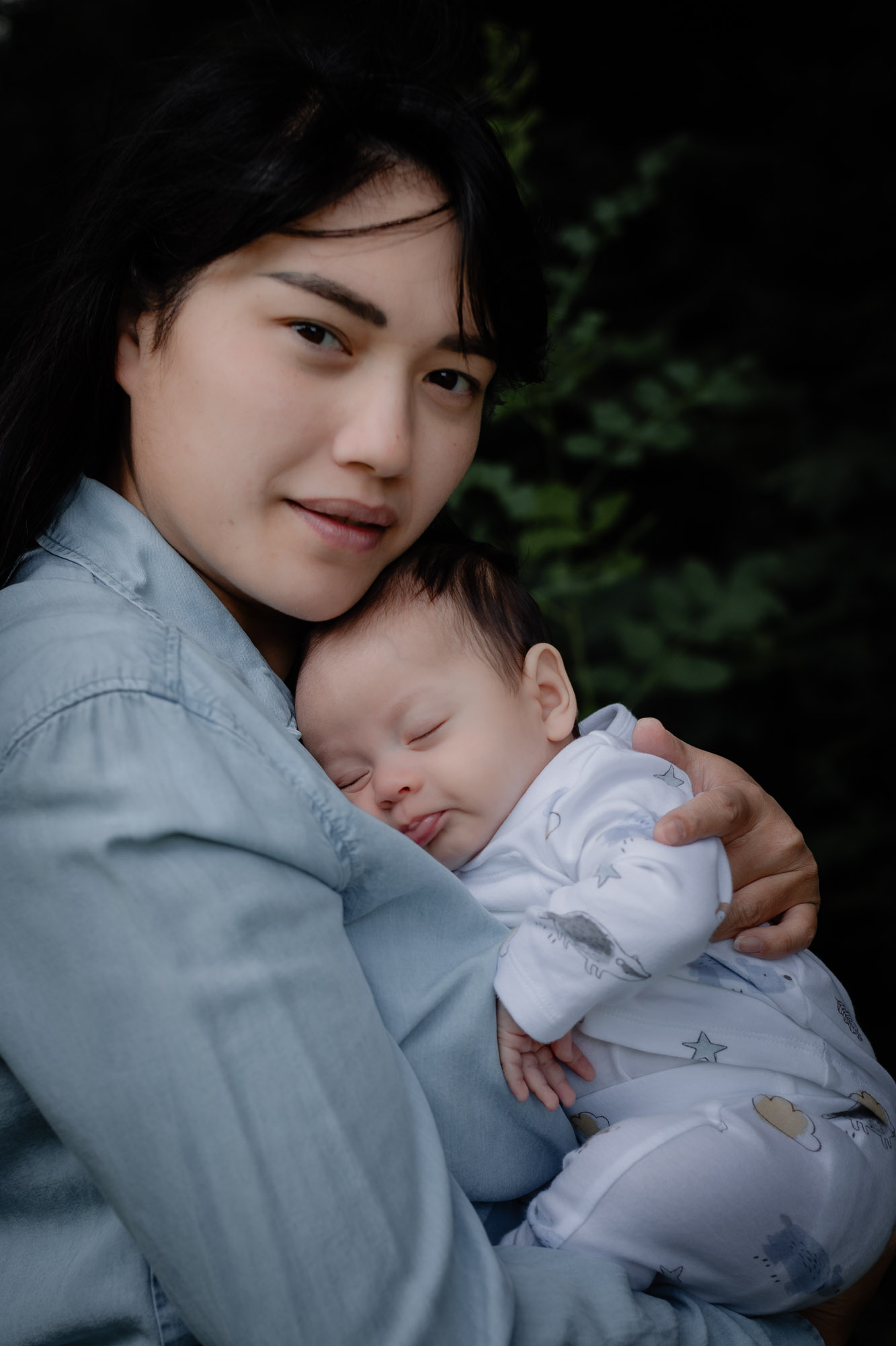 A young woman wearing a blue denim shirt cradling her baby son as he sleeps and she looks to camera on a baby photoshoot in Horsham West Sussex