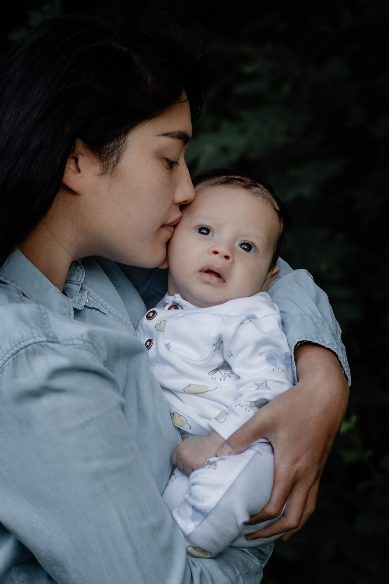 A young woman wearing a blue denim shirt cradling and kissing her newborn son as he looks towards the camera on a baby photoshoot in Horsham West Sussex