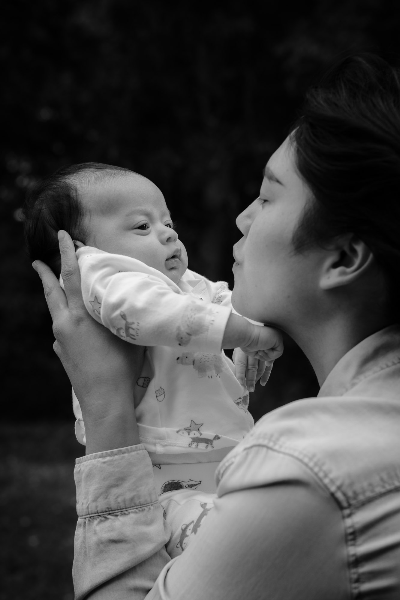 A black and white photo of a young woman holding her newborn out in front of her and making faces to him while he stares at her, with eyes wide open, on a baby photoshoot in Horsham West Sussex.