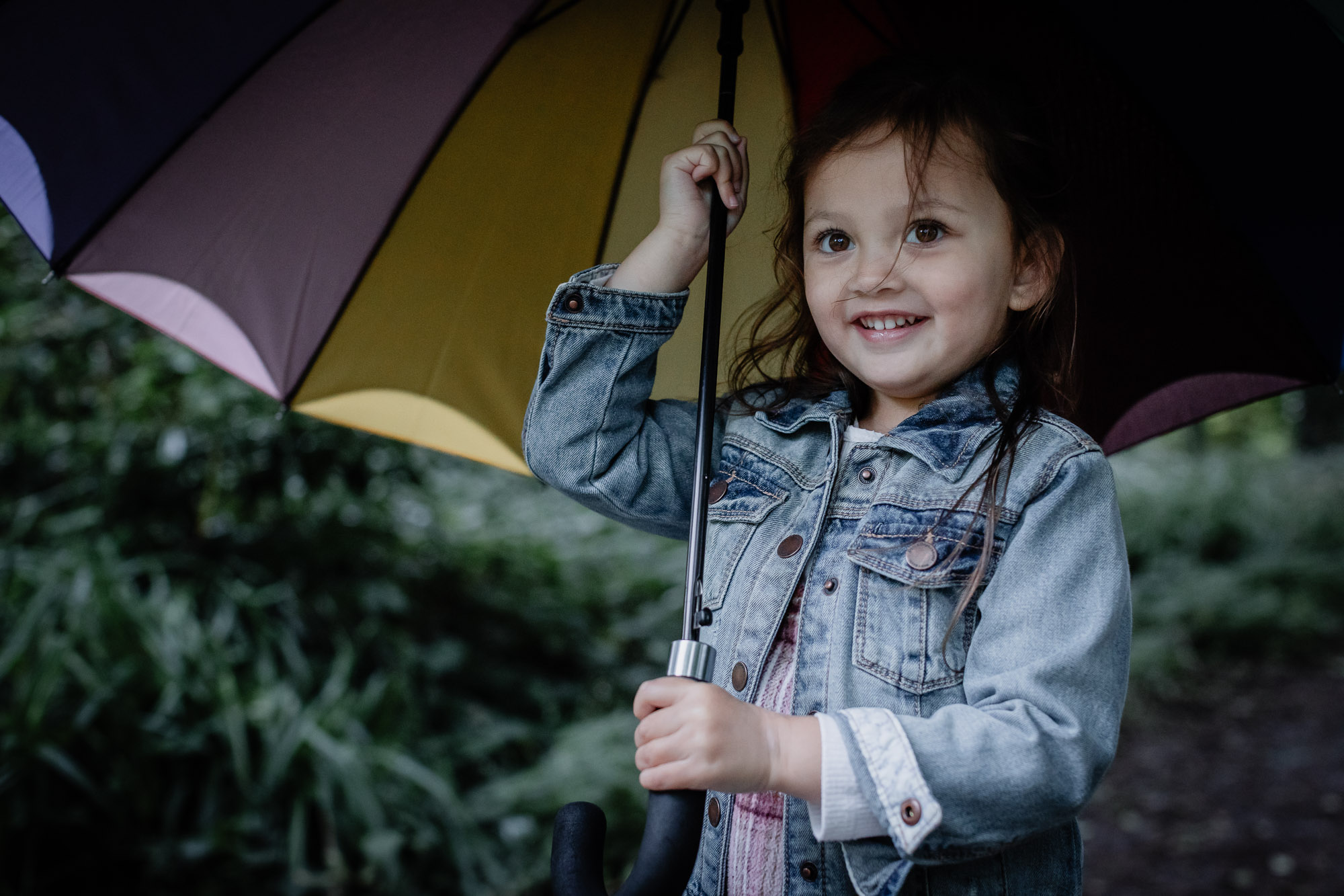 A little girl holding a multi-coloured umbrella over her head and smiling on a baby and family photoshoot in Horsham.