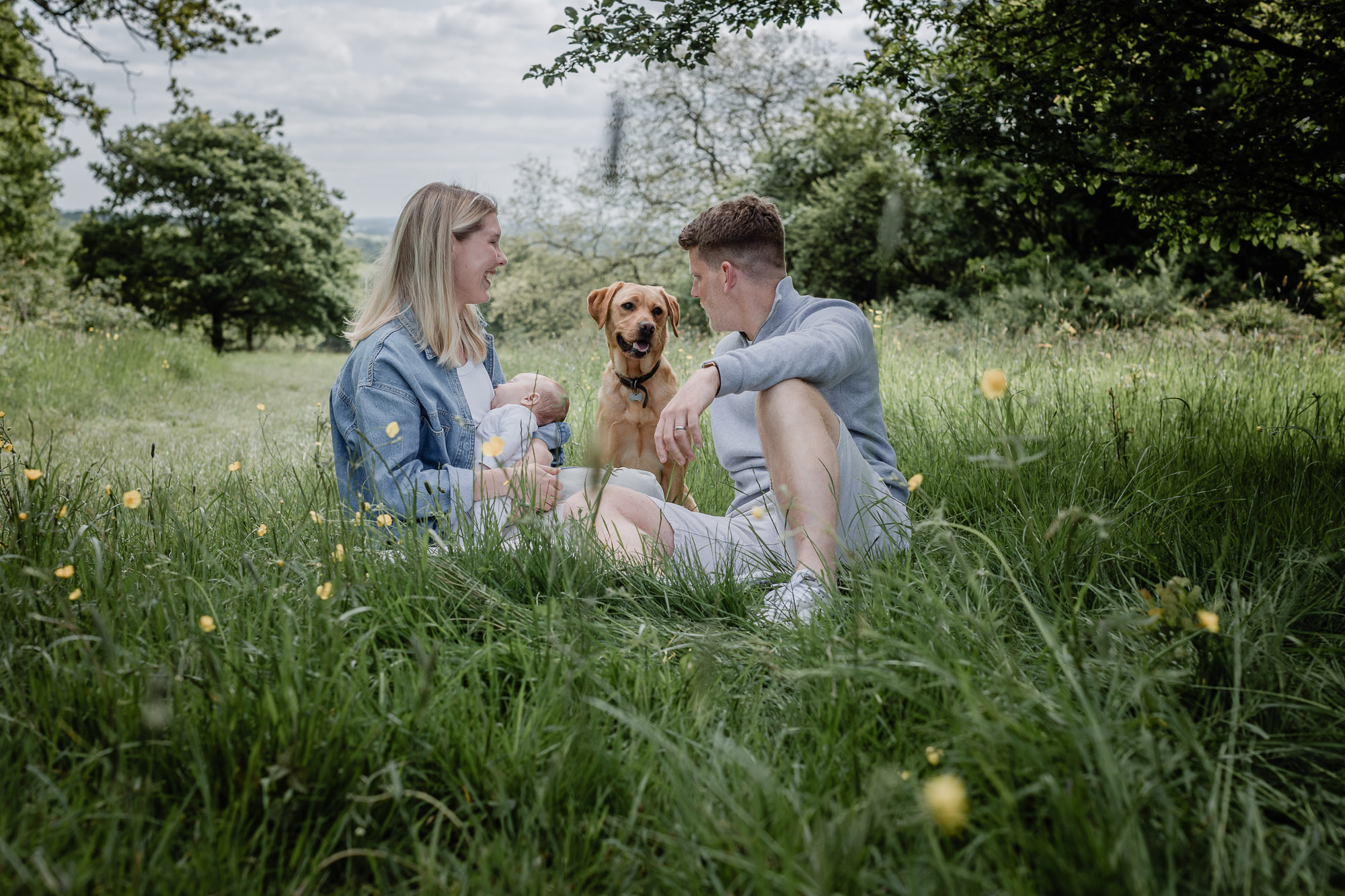 A young couple sitting on the grass in a field while their newborn is being breastfed and their red Labrador is sitting between them and looking intently at the father on an environmental newborn photoshoot in Horsham West Sussex.