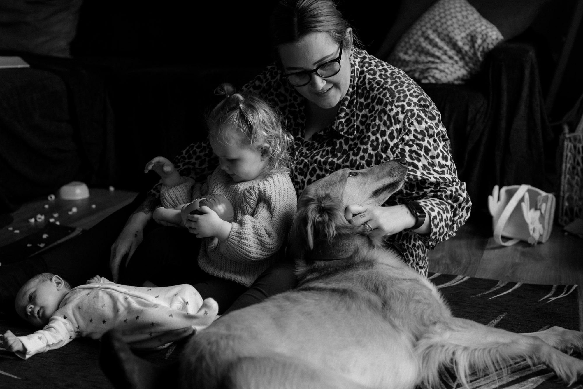 a family huddle of a young mother, with her 2 year old sitting in front of her and her newborn on the floor in front. their lurcher dog lies beside them and puts his head on the mother's lap while she strokes him on a newborn photoshoot in Horsham West Sussex